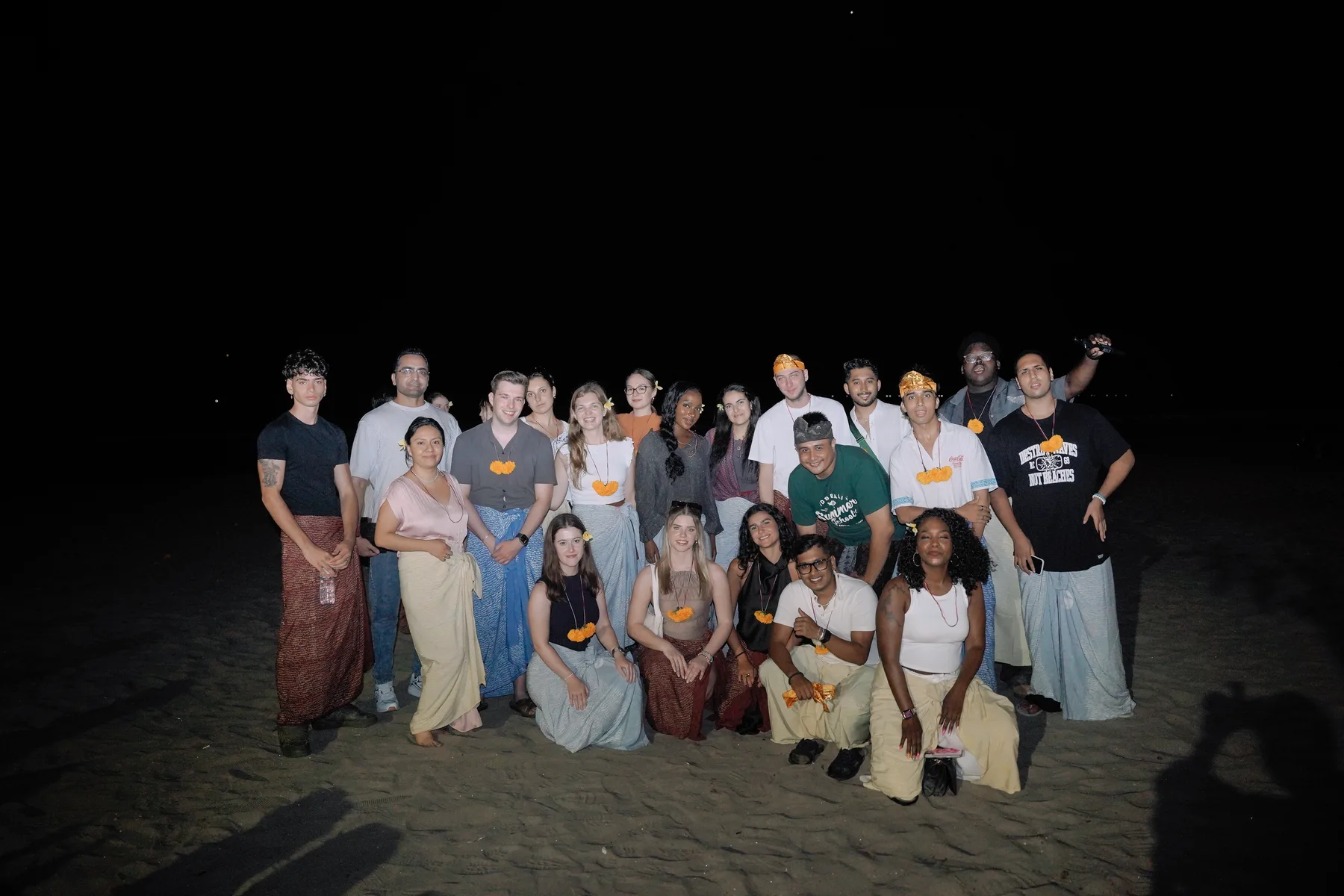 The Summer School 2026 cohort posing together on the beach at night, dressed in sarongs before the Welcoming Dinner