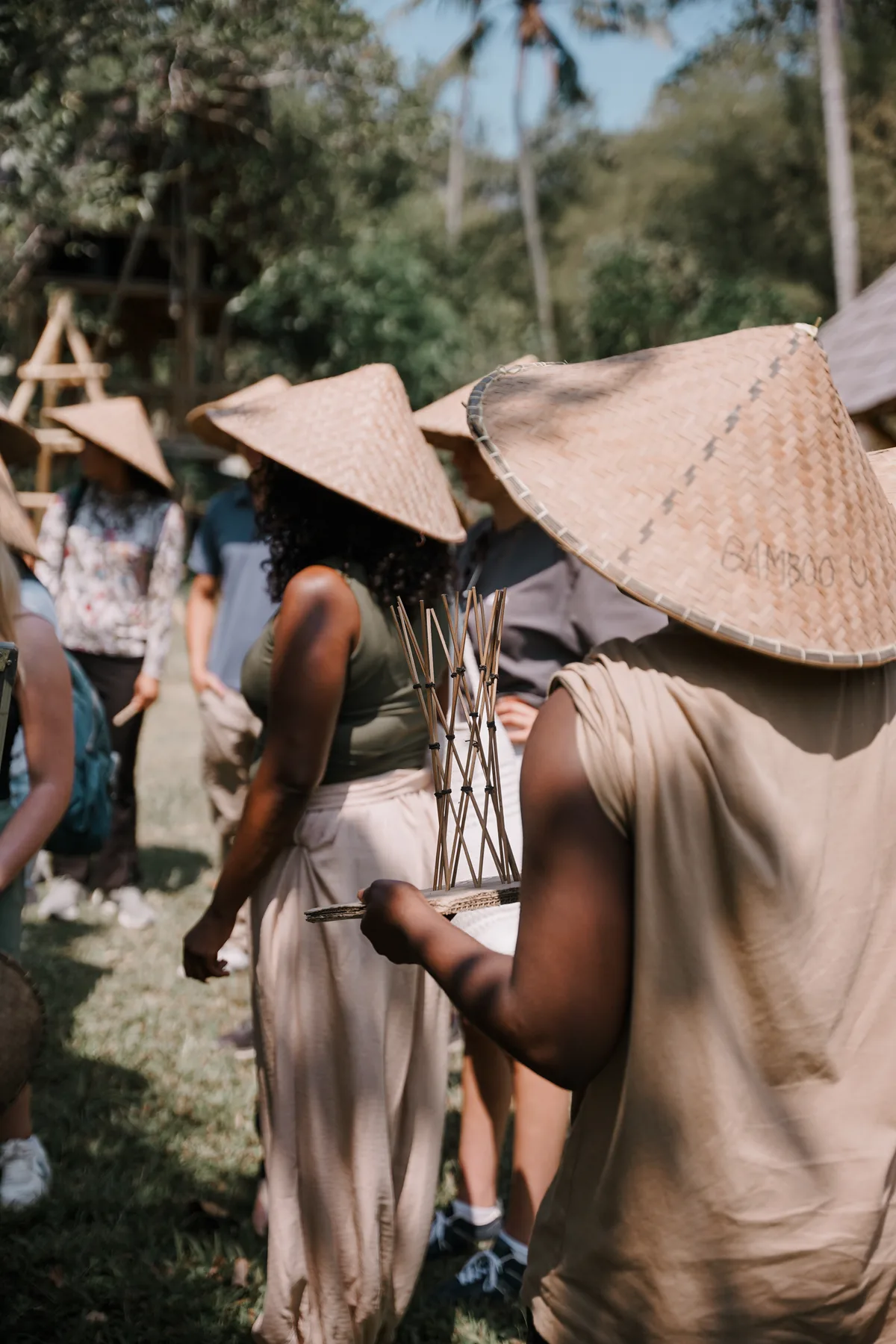 A student in a straw hat marked BambooU carrying a small lashed bamboo model