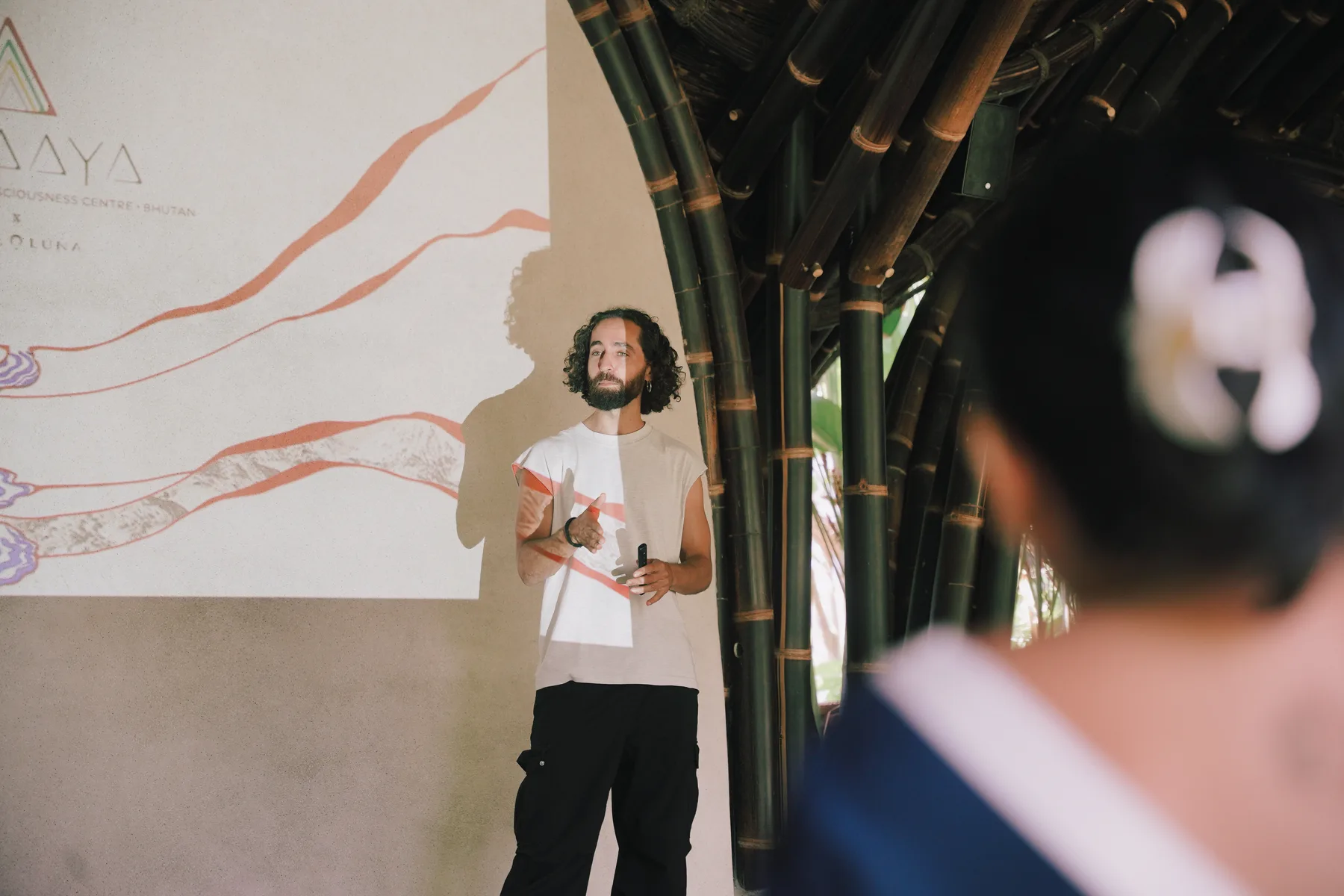 A studio principal presenting to a student under a bamboo roof structure, a projected slide visible on the wall