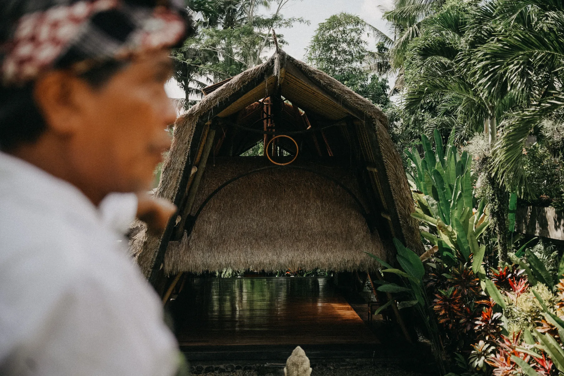 A traditional thatched-roof pavilion at Titi Ubud seen past a person in the foreground, surrounded by palm trees