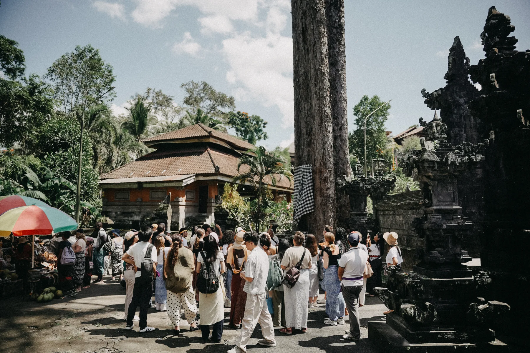 A wide view of the group gathered at the Goa Gajah temple complex, with traditional roofs and a market stall nearby