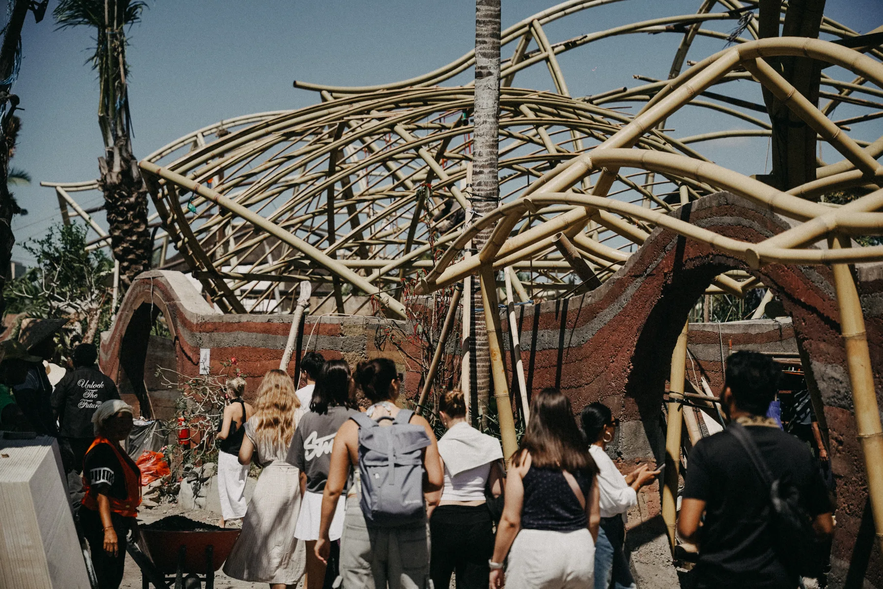 Students looking up at a large sculptural bamboo roof structure under construction at Nuanu, its curved beams woven overhead