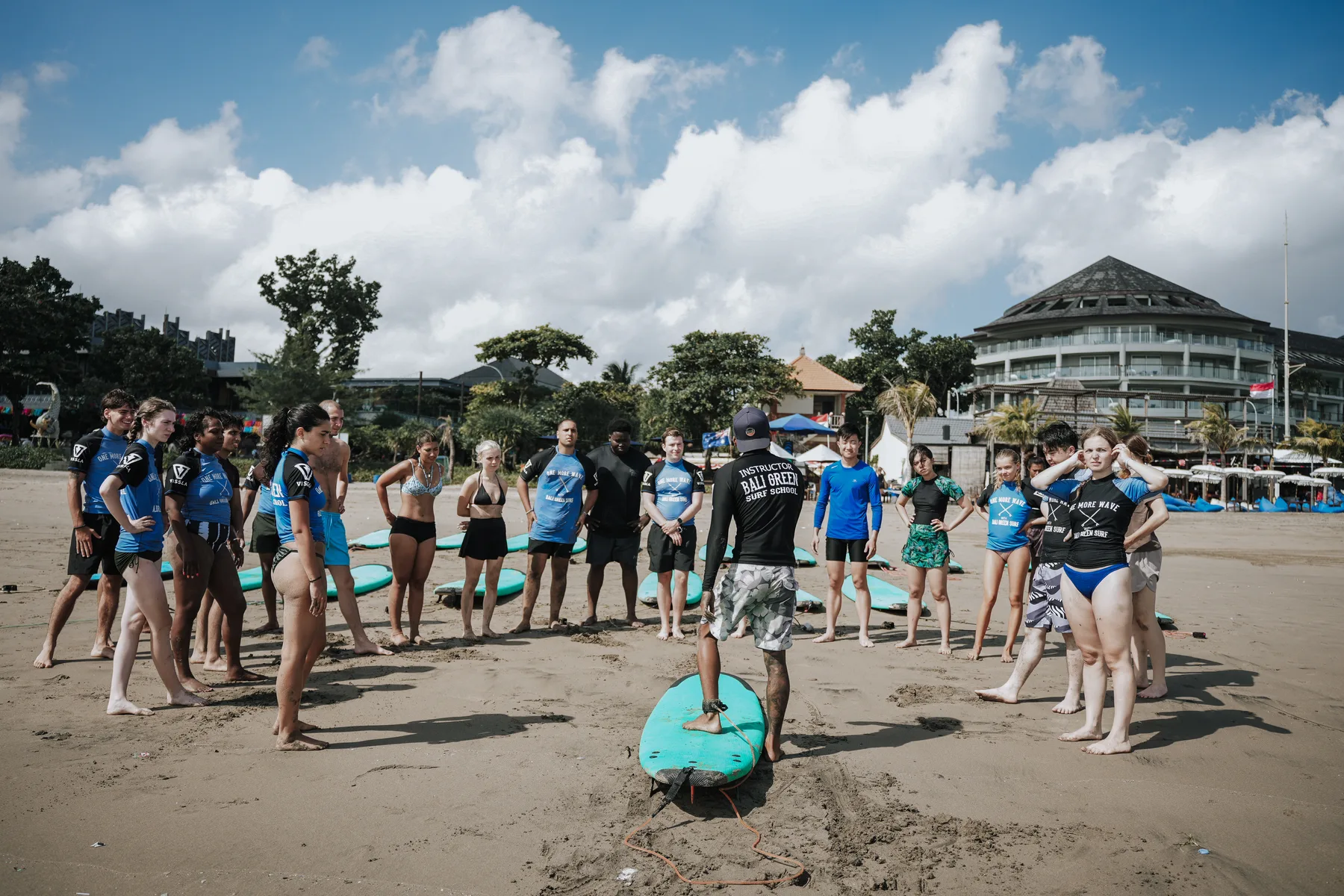 A surf instructor briefing a line of students standing beside their boards on the beach, hotels visible behind them