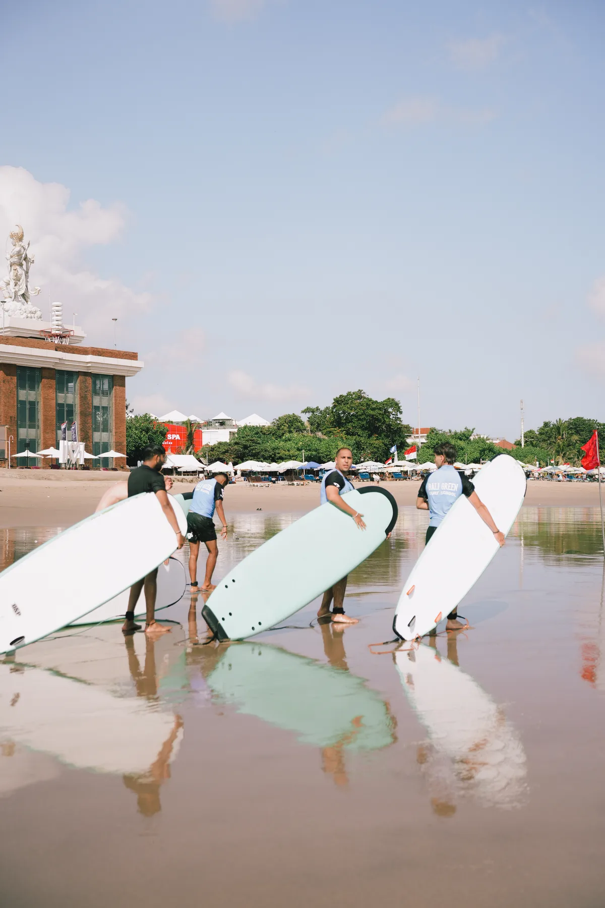 Students carrying surfboards toward the water, a landmark tower visible on the beach behind them