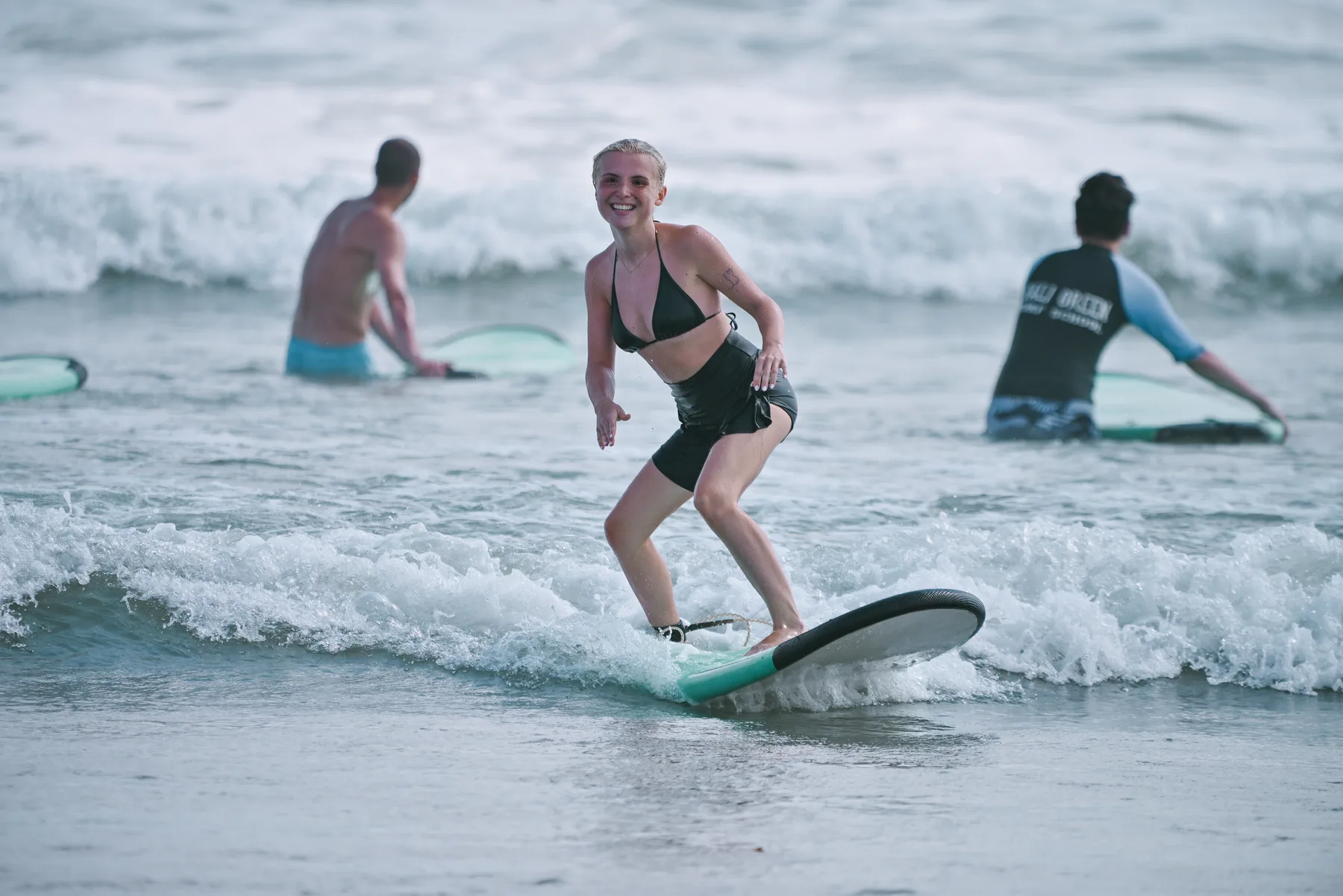 A student smiling while riding a small wave standing on a surfboard, two instructors watching from the water behind her