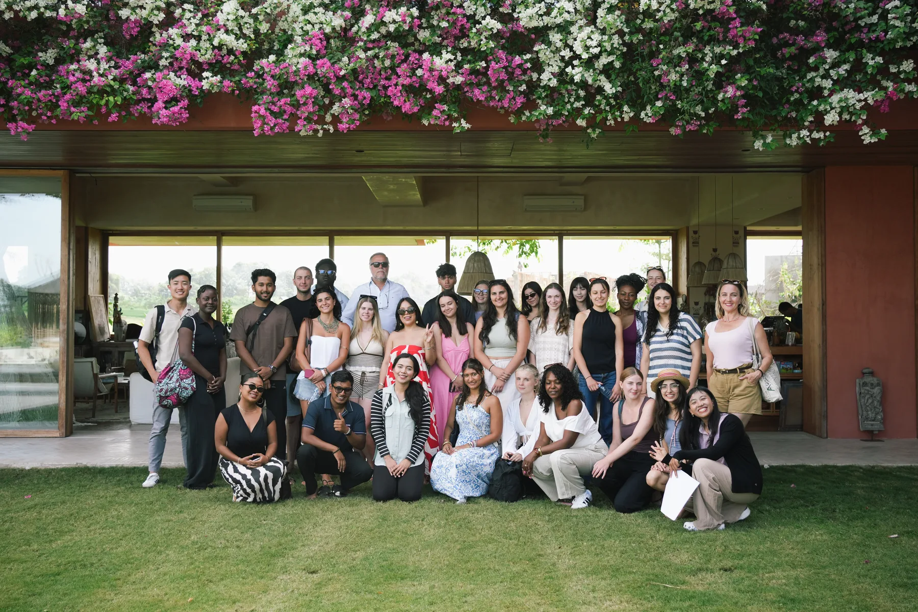 The full Summer School 2026 cohort posing on the lawn at Villa Nenek under a wall of pink and white bougainvillea