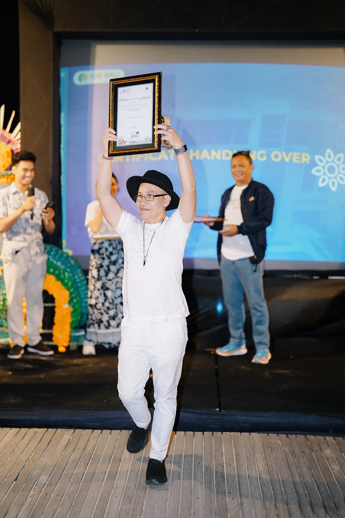 A lecturer holding a framed certificate above his head on stage during the certificate handover at the closing ceremony