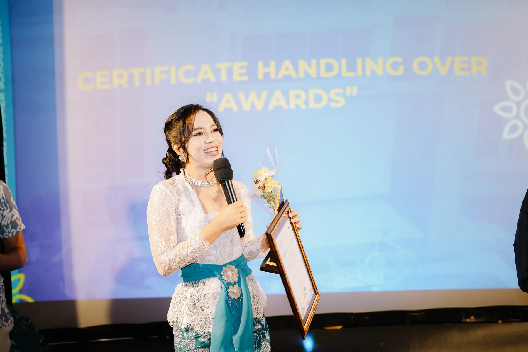 A student speaking into a microphone on stage, holding a framed certificate and flowers, with a screen reading Certificate Handling Over Awards behind her