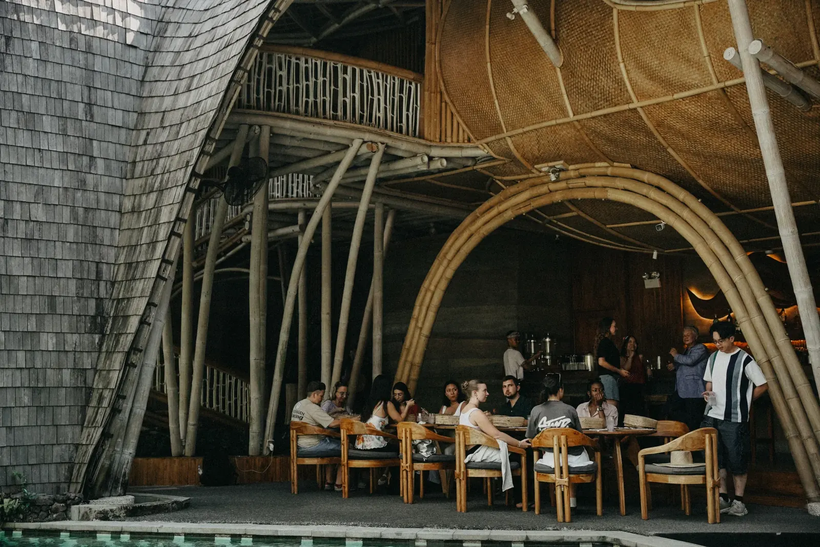Guests seated at wooden tables inside a bamboo-framed restaurant with arched beams and a shingled roof at Nuanu