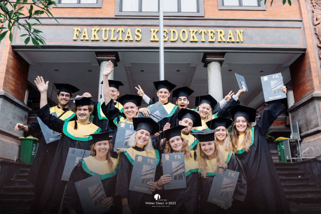 Graduating students from the Sports and Physiotherapy study abroad program at Universitas Udayana (UNUD) standing in front of the Medical Faculty building on graduation day, proudly holding their certificates.