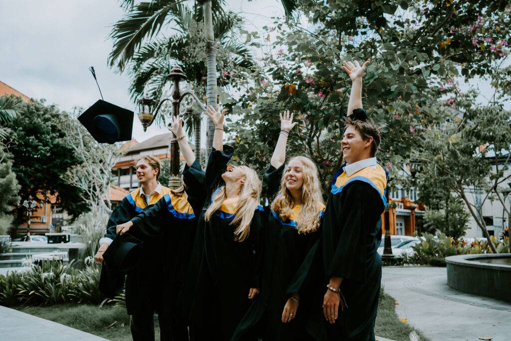 Graduates from the Southeast Asian Architecture and Southeast Asian Engineering study abroad programs at Universitas Udayana (UNUD) throwing their hats in celebration on campus outdoors.